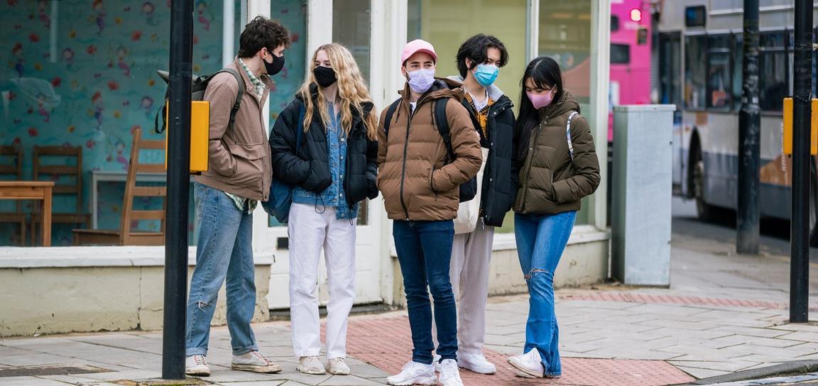 A group of five young adults wearing masks stands at a pedestrian crossing in an urban area.