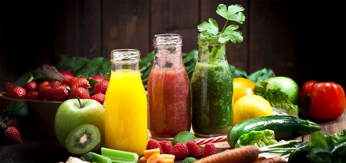 Three glass bottles of yellow, red, and green juices surrounded by various fruits and vegetables on a wooden table.