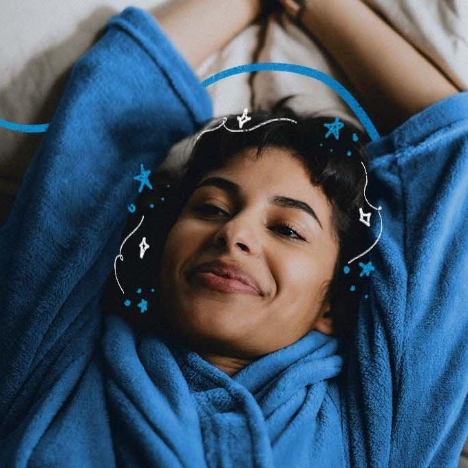 A smiling person lying on a bed wearing a blue dressing gown with their arms above their head