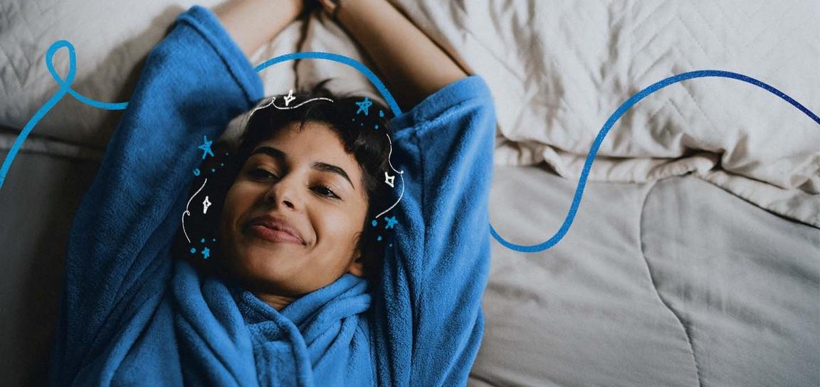 A smiling person lying on a bed wearing a blue dressing gown with their arms above their head