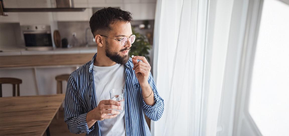 Person in a kitchen holding a glass of water and a pill near a window with white curtains.