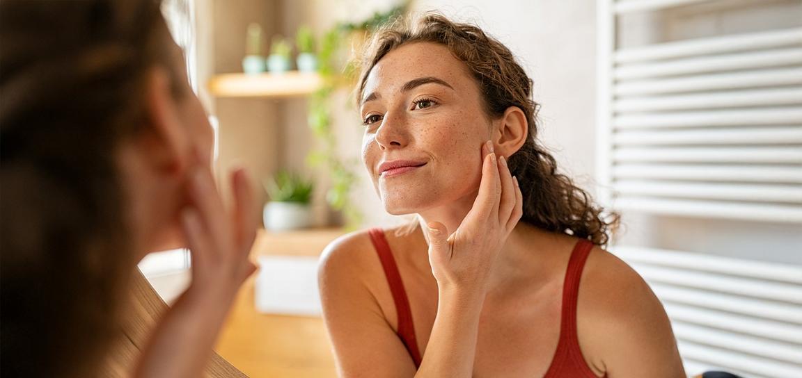 Woman examining her face in a mirror with a gentle smile.