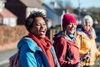 Three women walking and laughing outdoors on a sunny day in a residential area, wearing colourful jackets