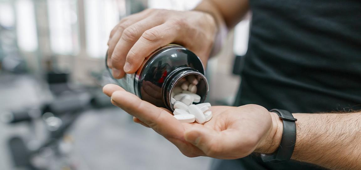 Hands pouring white tablets from a black bottle in a gym setting.