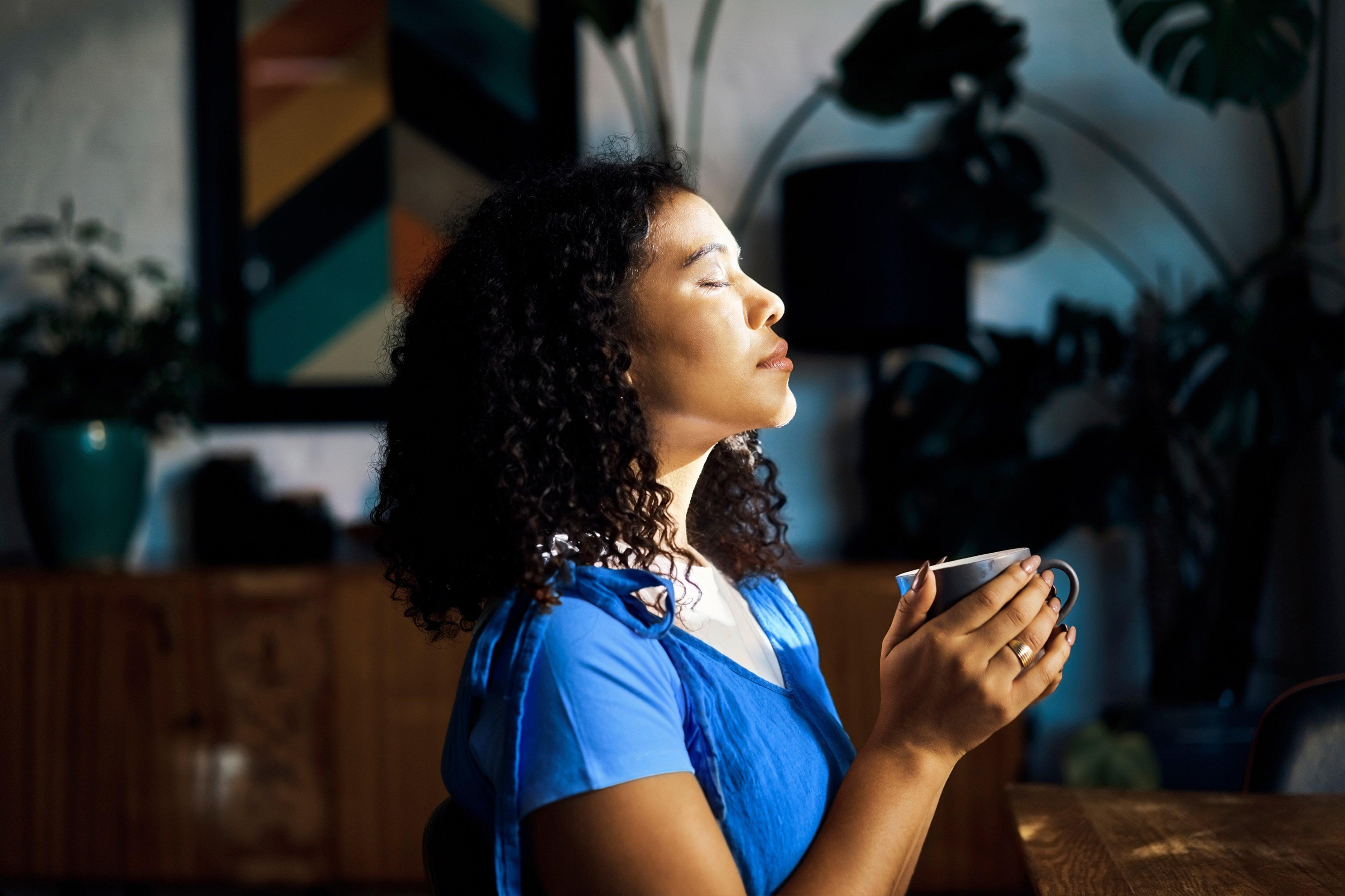 A woman stood with her eyes closed in front of a window, holding a mug