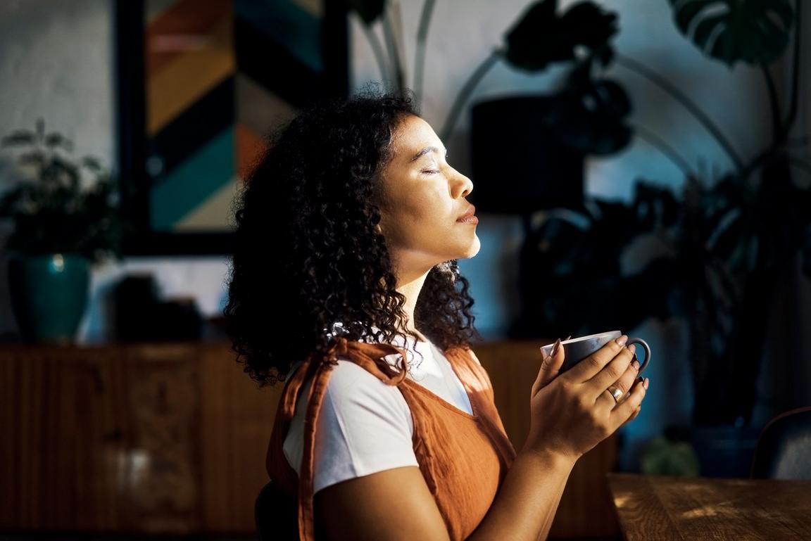A woman stood with her eyes closed in front of a window, holding a mug