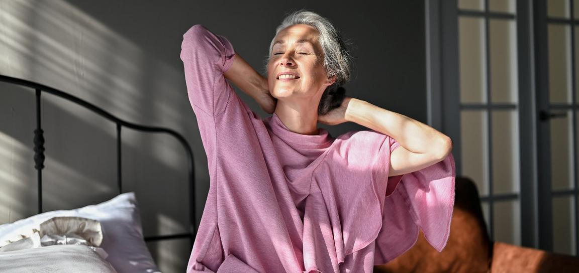 A person in a pink sweater sits on a bed, smiling with hands behind their head, in sunlight streaming through window blinds