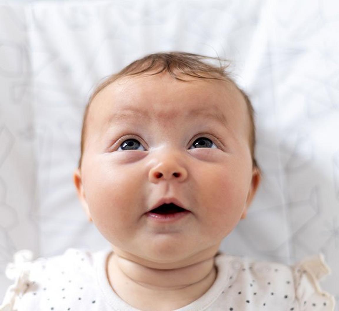 A baby with an inquisitive expression looks upward while lying on a patterned blanket.