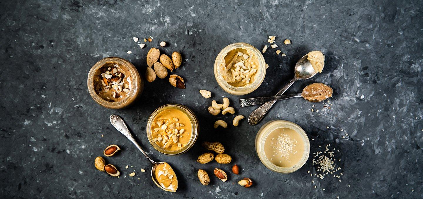 Assortment of nut and seed butters in jars surrounded by nuts, seeds and spoons.