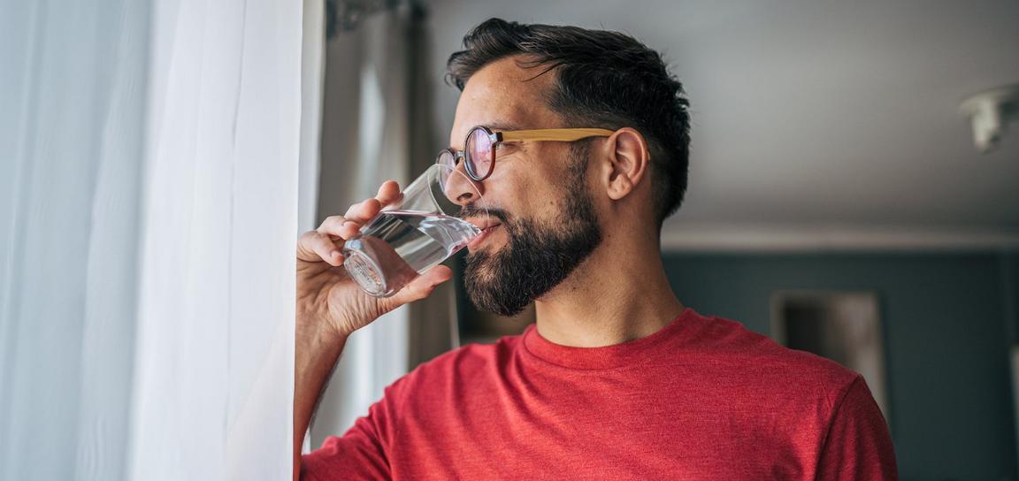 A man wearing glasses drinks water near a window with a sheer curtain.