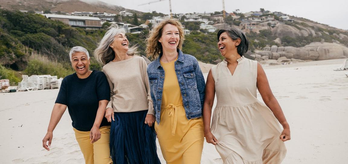 Four women walking and laughing on a beach with rocky hills and houses in the background.