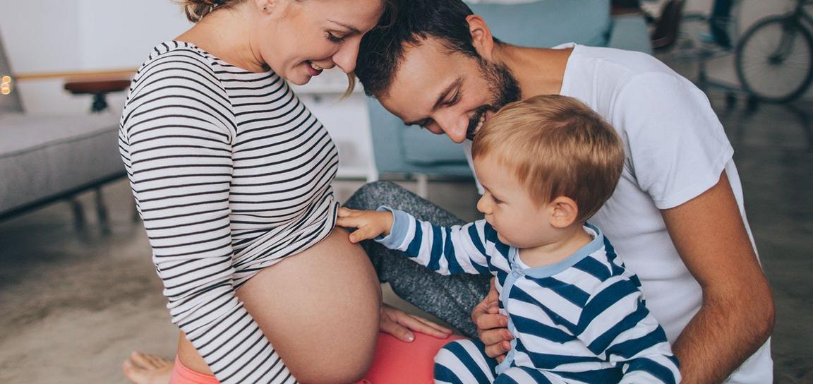 A pregnant woman sat on the floor with a man sat next to her and a baby placing their hand on the woman's stomach