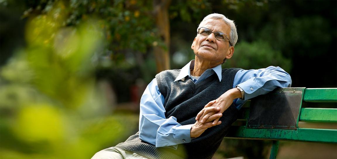 An elderly person sitting thoughtfully on a green bench outdoors, surrounded by greenery.