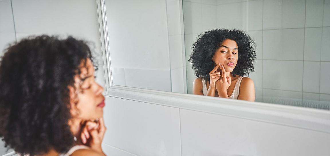 A young woman with curly hair examines her reflection in a bathroom mirror, touching her face thoughtfully.