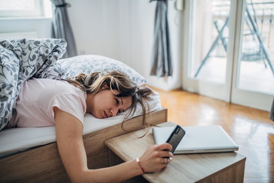 A woman lies in bed holding a smartphone above a small table with a closed laptop nearby in a bright bedroom.