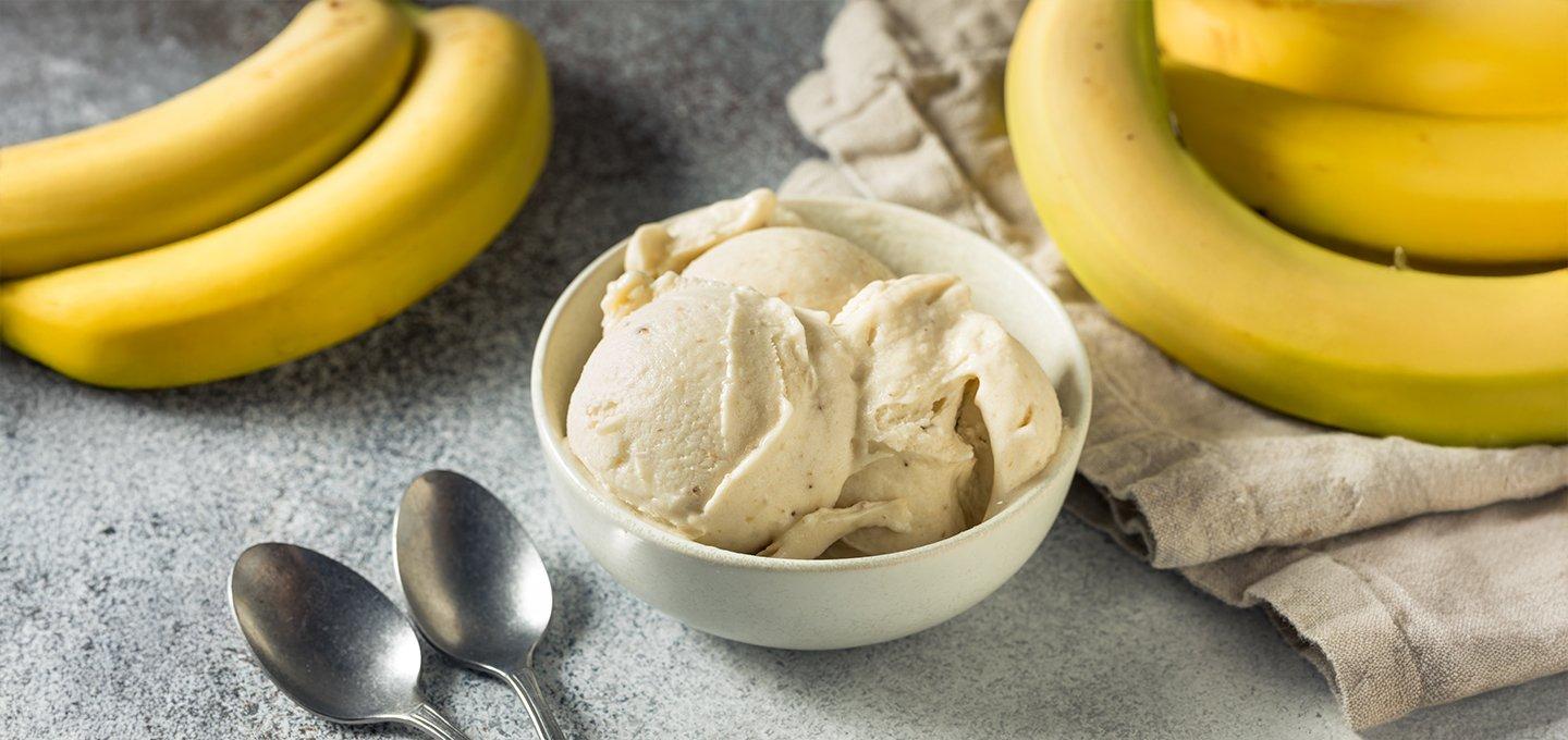 Bowl of banana ice cream with bunches of bananas and two spoons on a gray surface.