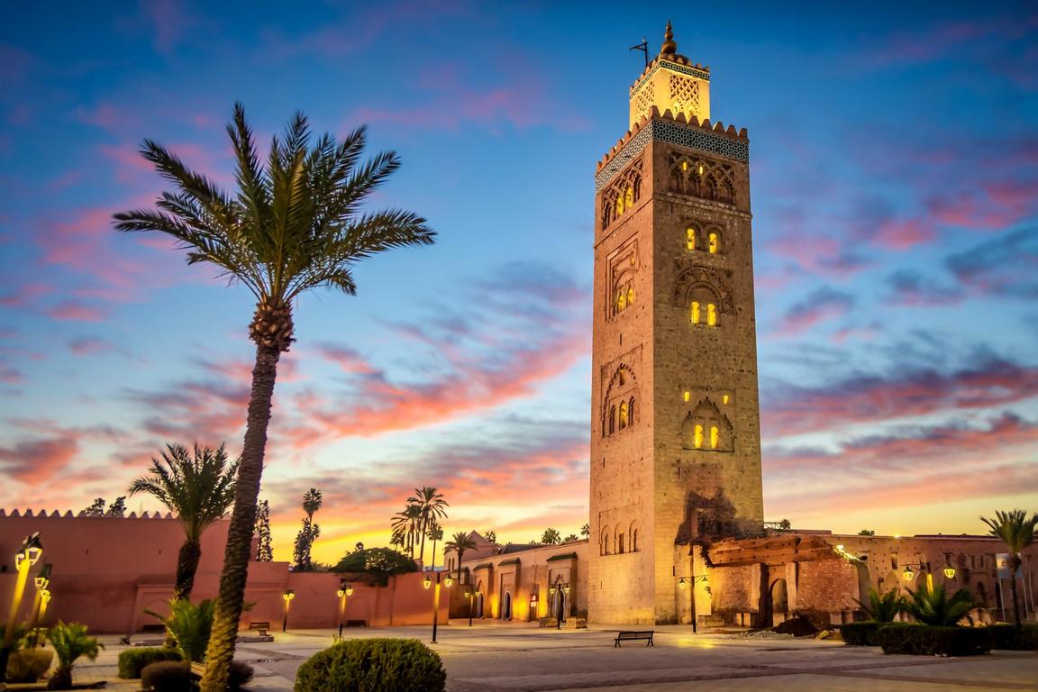 Tall illuminated stone tower at sunset with a palm tree and colorful sky.