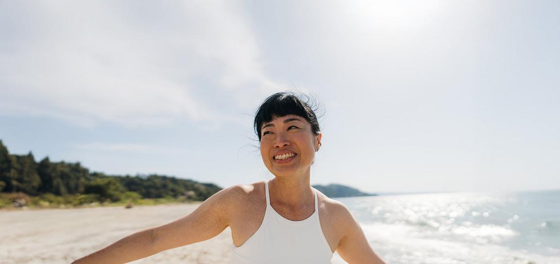 A person smiling on a sunny beach with the ocean and trees in the background.