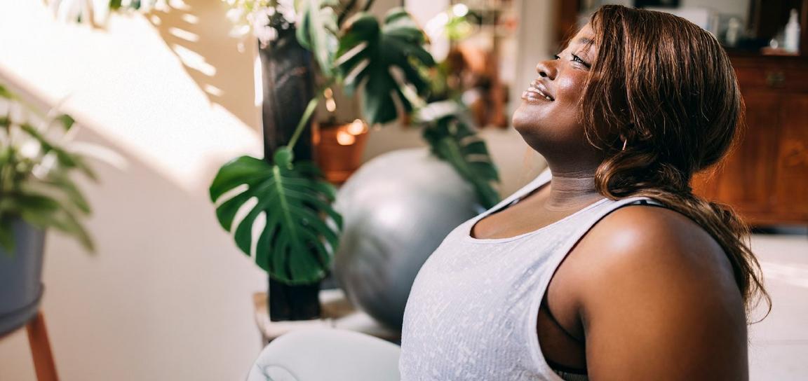 A woman in a gray tank top sitting indoors, surrounded by plants and soft sunlight.