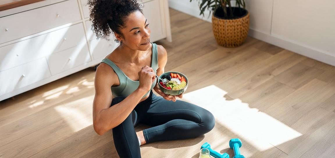 Woman eating from a bowl while sitting on the floor, with weights nearby in a sunlit room.