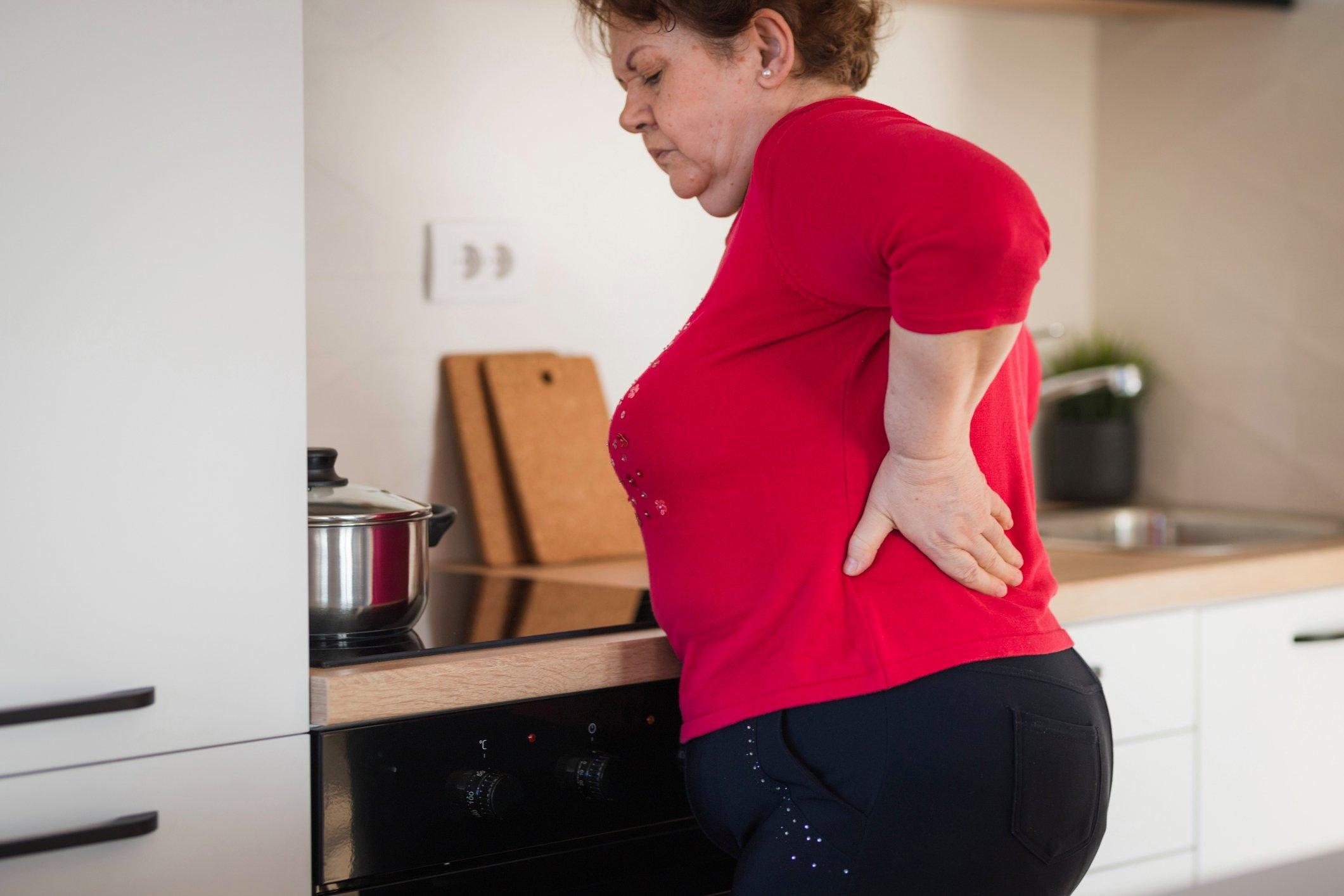Woman in a red shirt holding her back in a kitchen next to a stovetop.