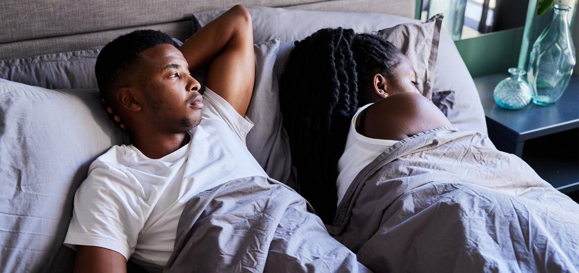 A couple in bed; the man is awake, looking up, while the woman sleeps, covered with a gray sheet.