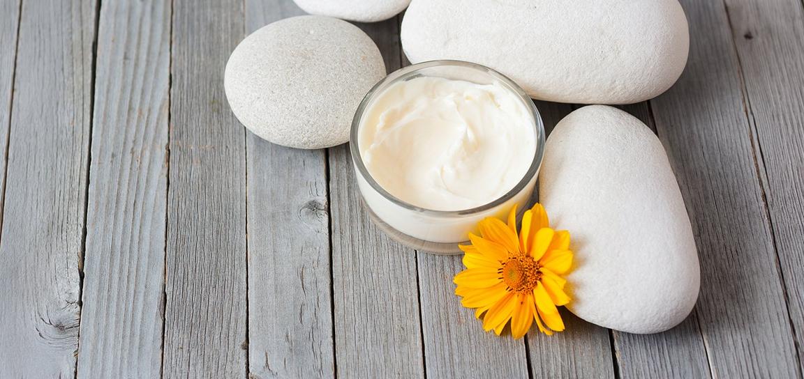 A glass jar of cream sits on gray wooden planks next to smooth light-colored pebbles and a yellow flower.