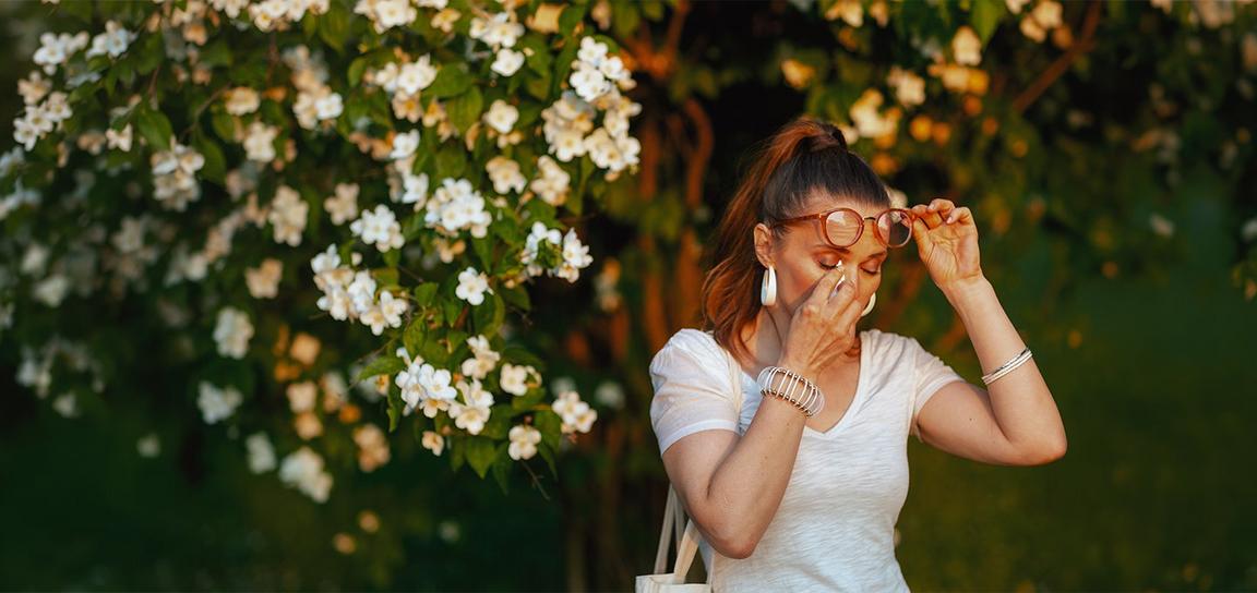 Person standing outdoors beside blooming white flowers, holding glasses and pinching the bridge of the nose, suggesting seasonal allergy or hay fever symptoms in a garden setting.