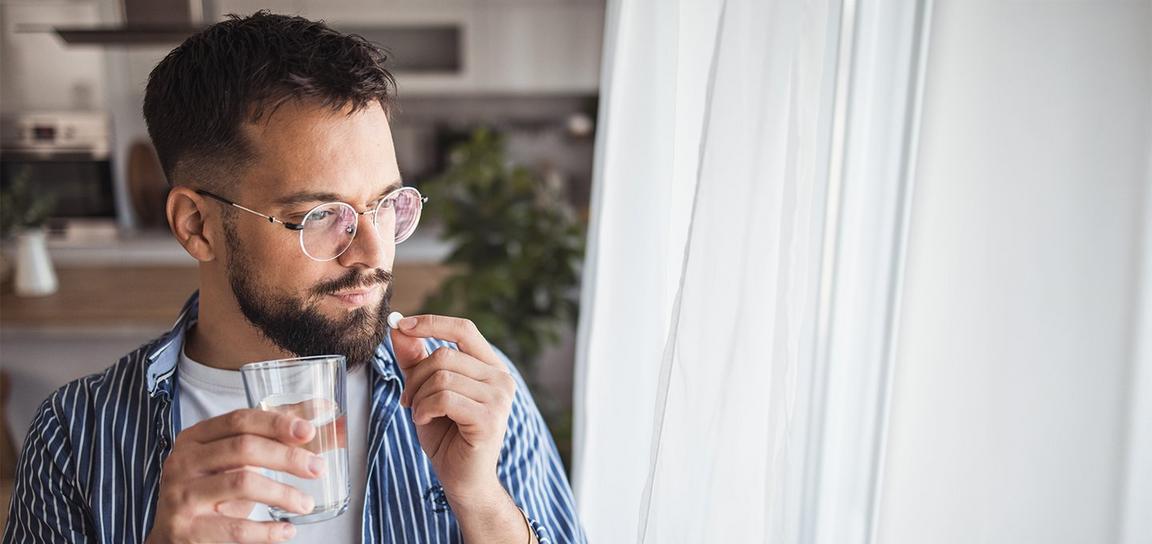A man holding a glass of water and a pill, looking out of a window.
